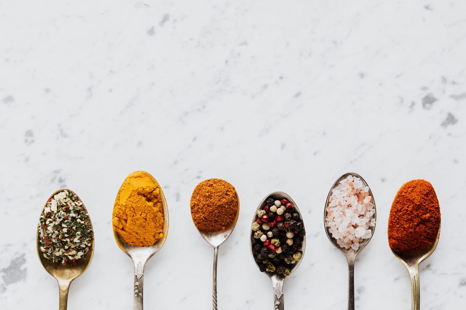 Six spoons of spices and seasonings on white marble: herbs, turmeric, curry powder, peppercorns, rock salt and paprika